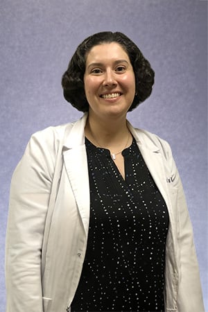 Portrait of Dr. Luba Kisilyuk, DDS wearing a white dental coat, smiling at the camera against a neutral background. She has dark, curly hair and is facing forward with a friendly expression.
